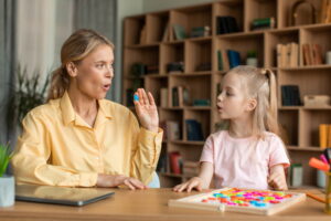Caring mother teaching small preschool child daughter pronouncing words and letters correctly Caring mother teaching small preschool child daughter pronouncing words and letters correctly. Smart girl doing vocal exercises with professional teacher, sitting at desk